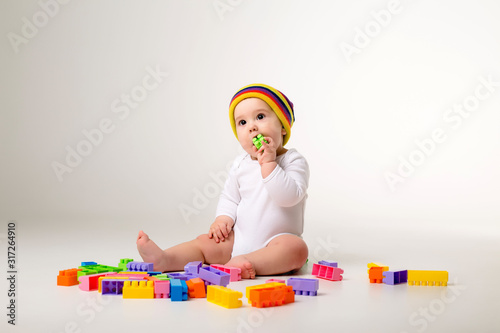 baby boy in white bodysuit and multicolored hat playing with a multicolored plastic constructor on a white background isolate, child development concept, space for text
