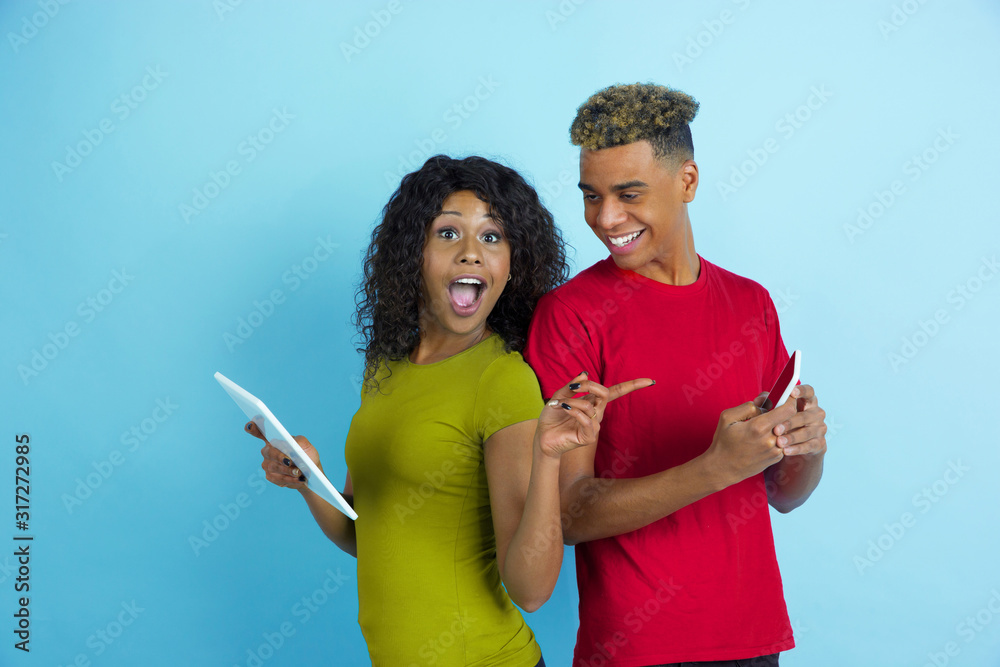 Using gadgets, laughting, pointing. Young emotional african-american man and woman in colorful clothes on blue background. Concept of human emotions, facial expession, relations, ad, friendship.