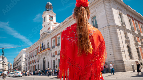 A young caucasian woman chulapa with red flower, traditional dress, and Spanish scarf at Puerta del Sol during San Isidro, the spring festival in May in the downtown of Madrid, the capital of Spain photo