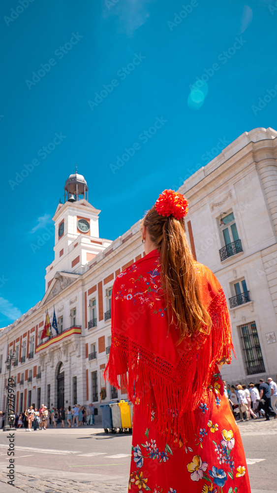 Fototapeta premium A young caucasian woman chulapa with red flower, traditional dress, and Spanish scarf at Puerta del Sol during San Isidro, the spring festival in May in the downtown of Madrid, the capital of Spain