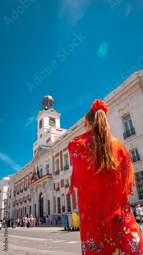 A young caucasian woman chulapa with red flower, traditional dress, and Spanish scarf at Puerta del Sol during San Isidro, the spring festival in May in the downtown of Madrid, the capital of Spain