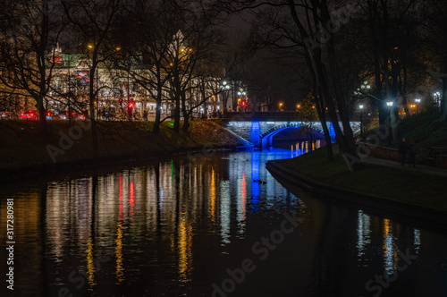 Small bridge lightened with blue light over Riga's City Canal by night with the city lights reflecting in the water in foggy November evening. National Theater in background