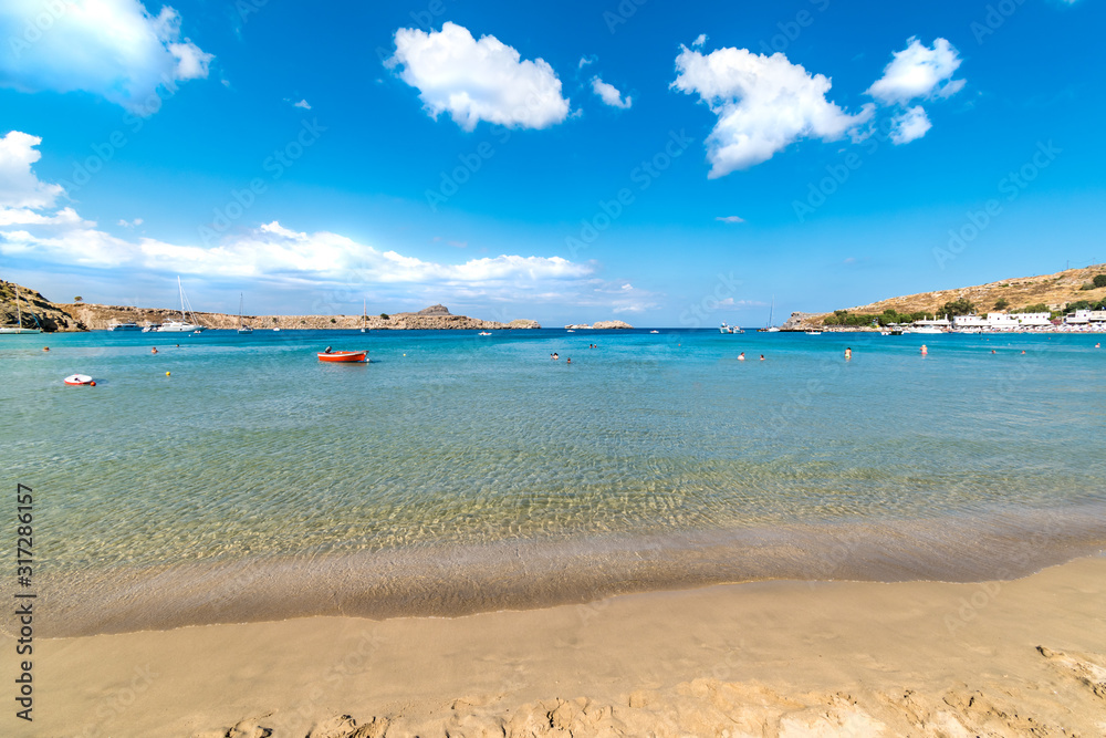 View of sandy beach and people in sea in Bay of Lindos, clouds on blue sky  (Rhodes, Greece)