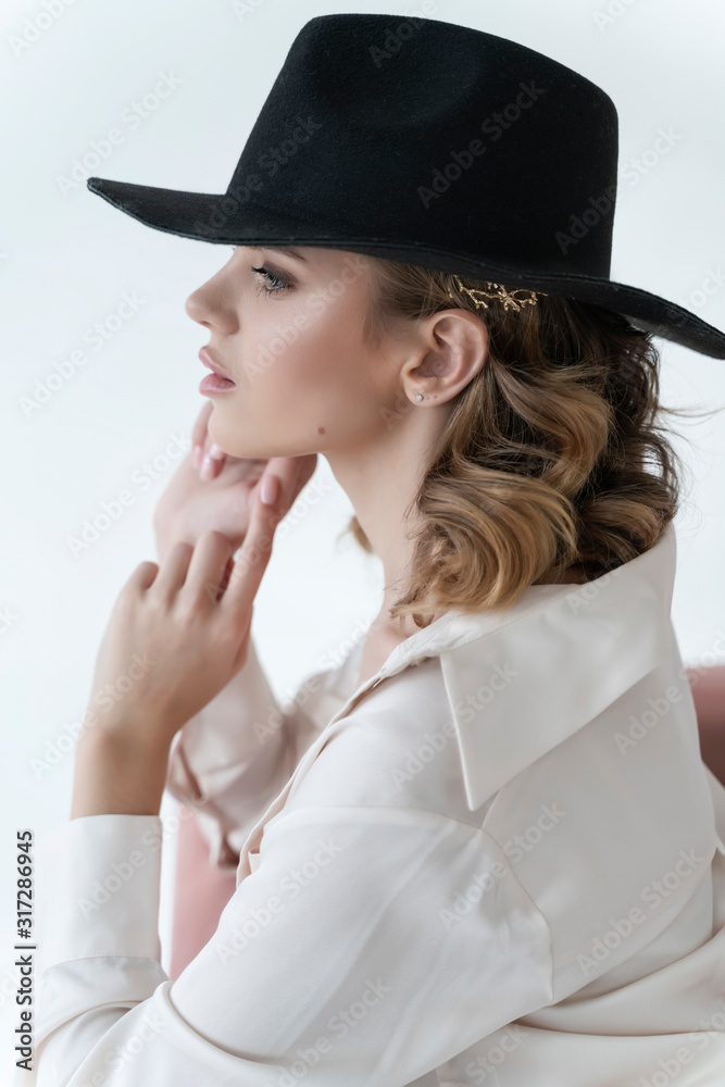 A beautiful girl wearing trousers, an unbuttoned shirt over a bra and a black hat sits thoughtfully and relaxed on a sofa on a white background. Fashionable, advertising and commercial design. A beautiful girl wearing trousers, an unbuttoned shirt over a bra and a black hat sits thoughtfully and relaxed on a sofa on a white background. Fashionable, advertising and commercial design.