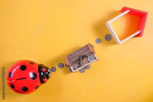 Ladybug piggy bank surrounded by coins and mini house on yellow background