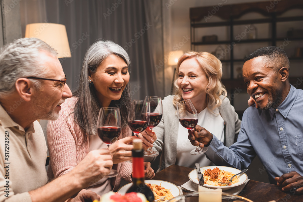 smiling multicultural friends talking and clinking with wine glasses during dinner smiling multicultural friends talking and clinking with wine glasses during dinner