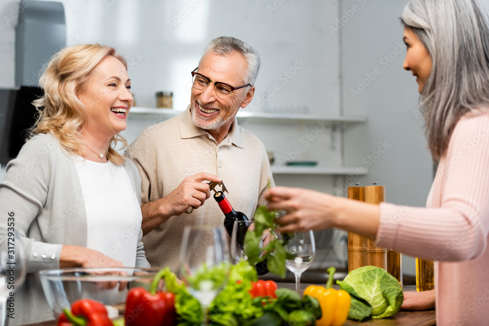 smiling woman cooking and talking with friend, man opening bottle with wine