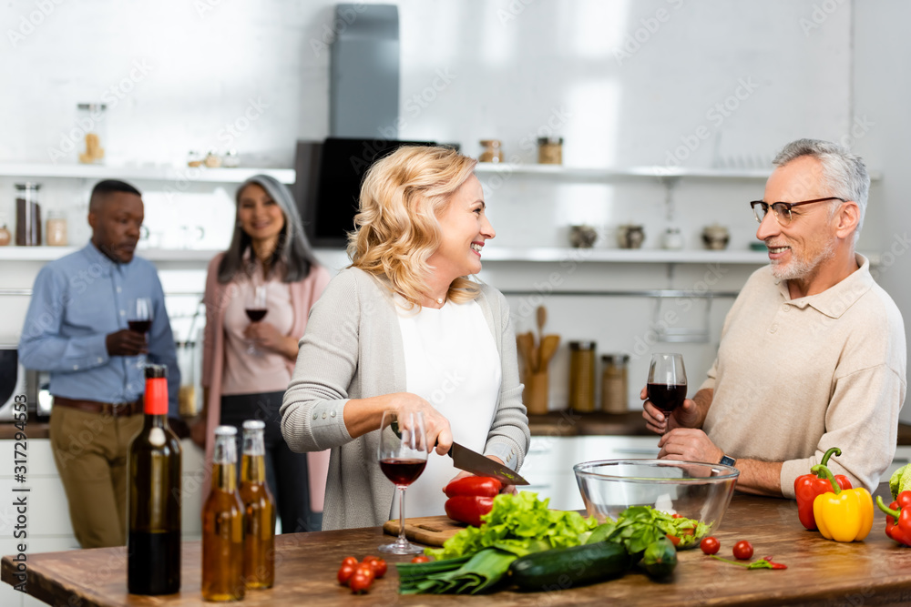 selective focus of woman cutting bell pepper and talking with man, multicultural friends talking on background