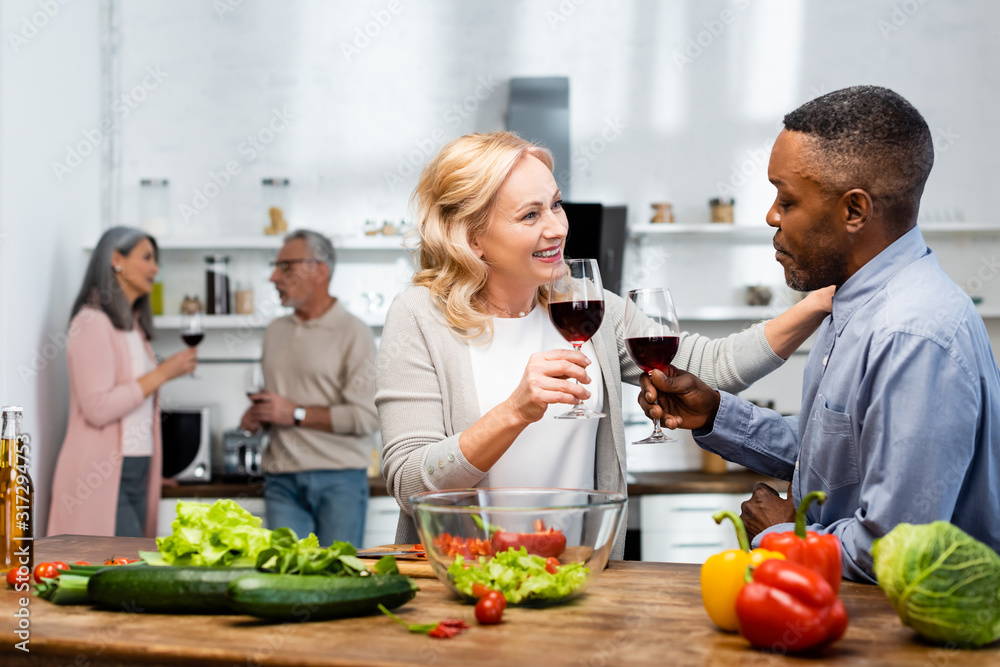 selective focus of smiling woman and african american man talking and holding wine glasses, multicultural friends talking on background