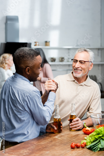 african american man holding hands and talking with smiling friend in kitchen