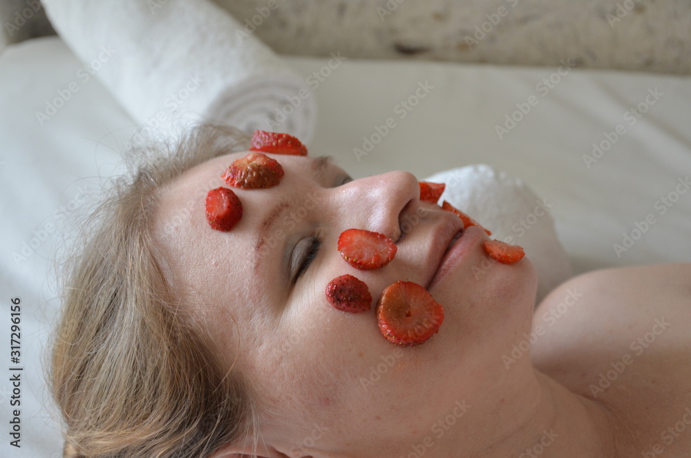 Fototapeta premium Young beautiful woman getting cosmetic fruit facial mask of fruit strawberry at beauty salon on a white towel indoors. natural cosmetic