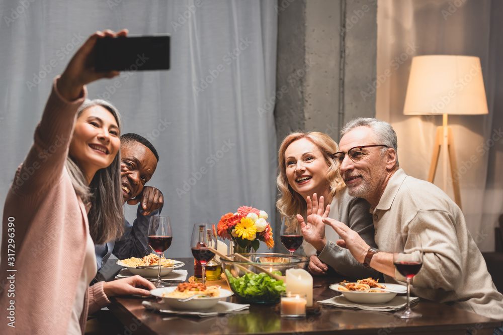 smiling multicultural friends taking selfie with smartphone during dinner smiling multicultural friends taking selfie with smartphone during dinner
