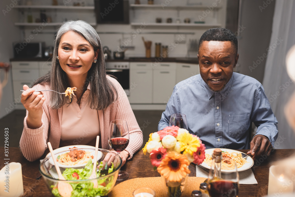 selective focus of smiling multicultural man and woman talking and eating pasta during dinner