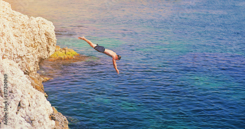Fotografie Young man jumping off cliff into blue water ocean at sunset