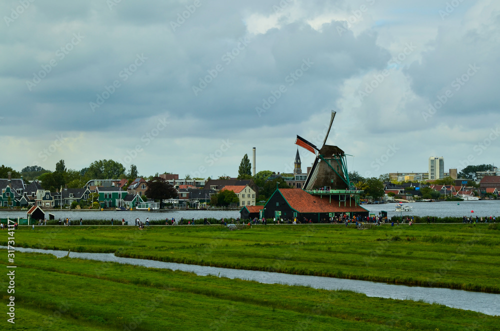 Fototapeta premium Zaanse Schans, Holland, August 2019. North-east of Amsterdam is a small community located on the quay of the Zaan river. View from above of the fields with mills, tourists are noticed. Cloudy day.