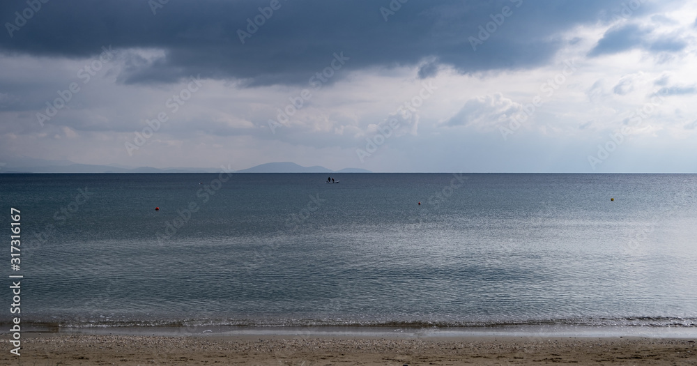 Fluffy grey-blue clouds meet calm dark blue sea far from the sandy coast. Mountains background, space.