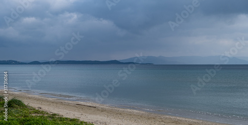 Fluffy grey-blue clouds meet calm dark blue sea far from the sandy coast. Mountains background  space.