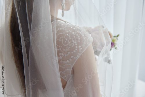 Portrait of beautiful bride in wedding dress standing with bridal bouquet near window in room, copy space. Bride's morning preparation. Wedding concept