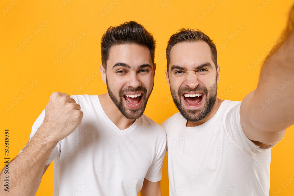 Close up of joyful young men guys friends in white t-shirts isolated on yellow orange background. People lifestyle concept. Mock up copy space. Doing selfie shot on mobile phone doing winner gesture. Close up of joyful young men guys friends in white t-shirts isolated on yellow orange background. People lifestyle concept. Mock up copy space. Doing selfie shot on mobile phone doing winner gesture.