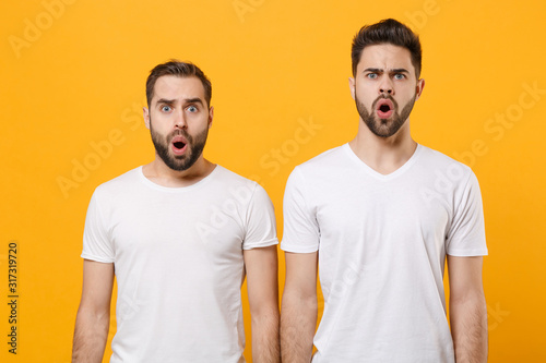 Shocked young men guys friends in white blank empty t-shirts posing isolated on yellow orange background studio portrait. People emotions lifestyle concept. Mock up copy space. Keeping mouth open.