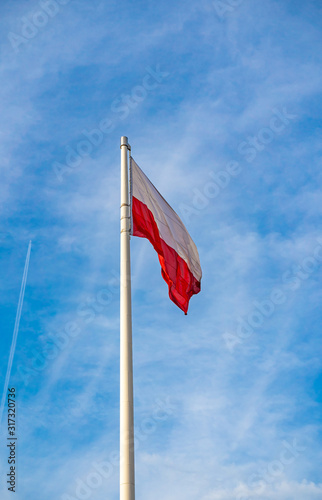 Polish majestic flag hanging on white pole and slowly waving in the wind against blue and cloudy sky.
