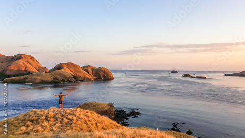 A man standing on top of a small island, enjoying the morning sun over Komodo National Park, Flores, Indonesia. Golden hour over the islands and sea. Some boats anchored to the bay. New day beginning photo
