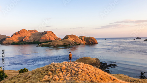 A man standing on top of a small island, enjoying the morning sun over Komodo National Park, Flores, Indonesia. Golden hour over the islands and sea. Some boats anchored to the bay. New day beginning photo