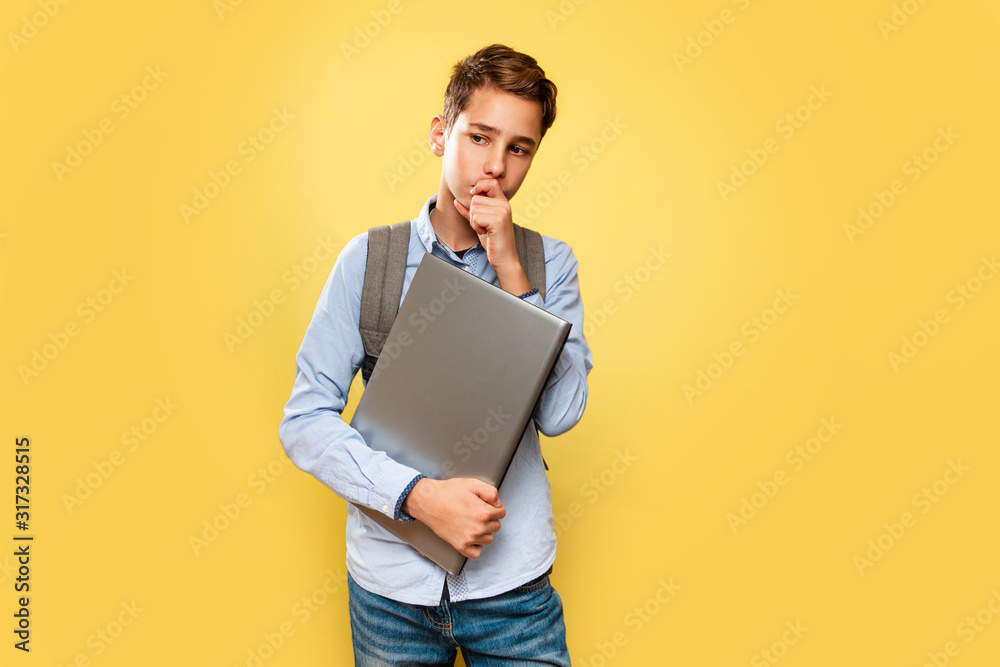 Concept of modern technologies and education. A young Caucasian teenage boy in a blue shirt and backpack, with a laptop in his hands, thoughtfully holding his chin. Yellow background. Copy space Concept of modern technologies and education. A young Caucasian teenage boy in a blue shirt and backpack, with a laptop in his hands, thoughtfully holding his chin. Yellow background. Copy space