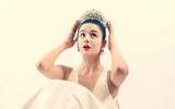 Closeup shot of pretty lady with natural makeup, wearing white dress, earrings and silver tiara with white gems. The girl with brown hair is posing on a light background