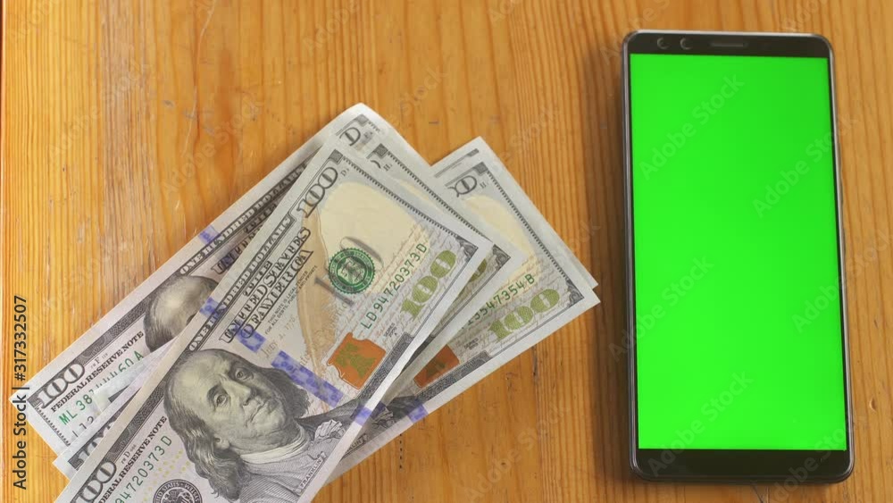 Hundred dollar bills fall next to phone greenscreen touchpad on the table. Mock-up with green screen cellphone. Money cash chroma key concept. Technology and economy. Personal budget. Home finance. Hundred dollar bills fall next to phone greenscreen touchpad on the table. Mock-up with green screen cellphone. Money cash chroma key concept. Technology and economy. Personal budget. Home finance.