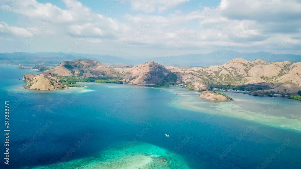 Fototapeta premium A drone shot of a paradise island with some boats anchored around in Komodo National Park, Flores, Indonesia. Brownish island turns into white sand beach and further into turquoise and navy sea.