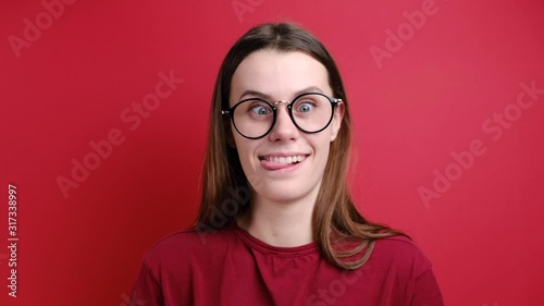 Cute young girl in glasses makes funny face, crosses eyes and sticks out tongue, plays fool, doesnt want to be responsible and looking at camera, stands over red background. Positive emotions photo