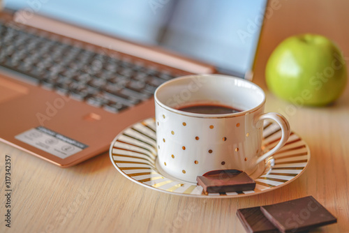 Tempting cup of coffee on the wooden table with laptop and apple on background. Piece of dark chocolate is on the saucer, break time in the office for refreshment, closeup