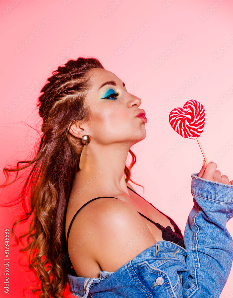 Beautiful brunette girl with blue makeup, pigtails and a sleeveless denim vest posing on a red background with a heart-shaped lollipop d