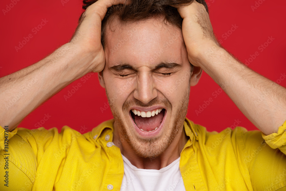 crazy man in yellow shirt holding head and screaming isolated on red background