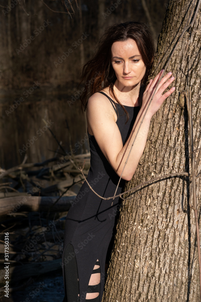 Φωτογραφία Young woman with dark long hair in black robes is leaning against the tree in the middle of the forest
