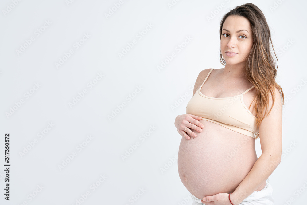 Attractive young pregnant woman in beige bra holds her hands on her swollen belly and looking at the camera, isolated over white background Attractive young pregnant woman in beige bra holds her hands on her swollen belly and looking at the camera, isolated over white background