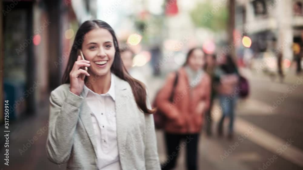 Attractive and cheerful businesswoman using smartphone in a crowded street. She is talking on the phone and laughing while walking. City at sunset. Attractive and cheerful businesswoman using smartphone in a crowded street. She is talking on the phone and laughing while walking. City at sunset.