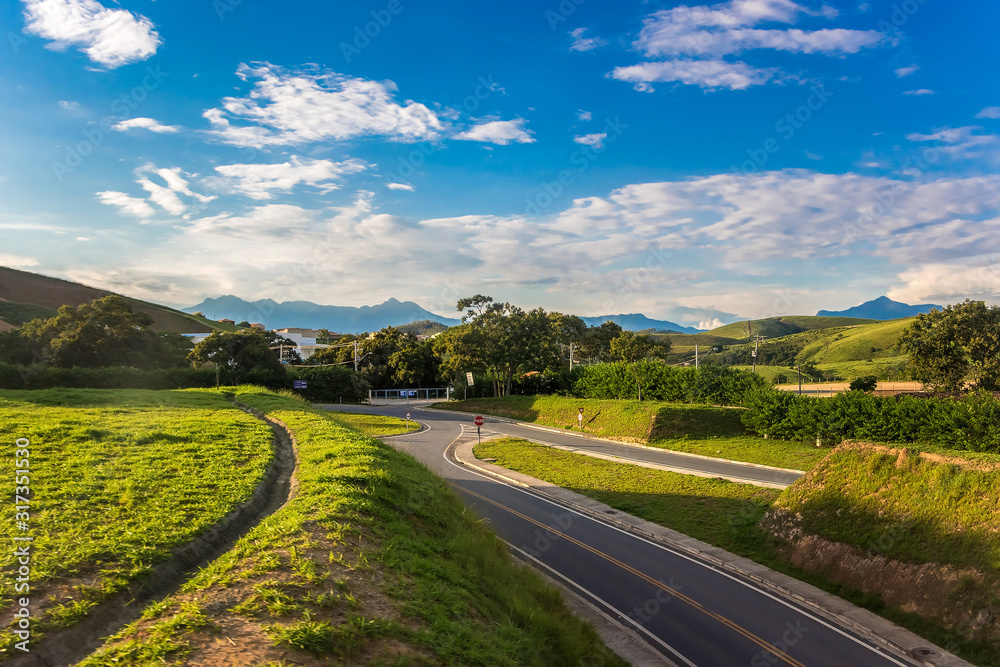 Naklejka premium Rural Road Among Grean Fields. Amazing Blue Sky With Beautiful Clouds, Mountains, Meadow With Fresh Green Grass. Highway In Rio de Janeiro District. Brazilian Countryside.