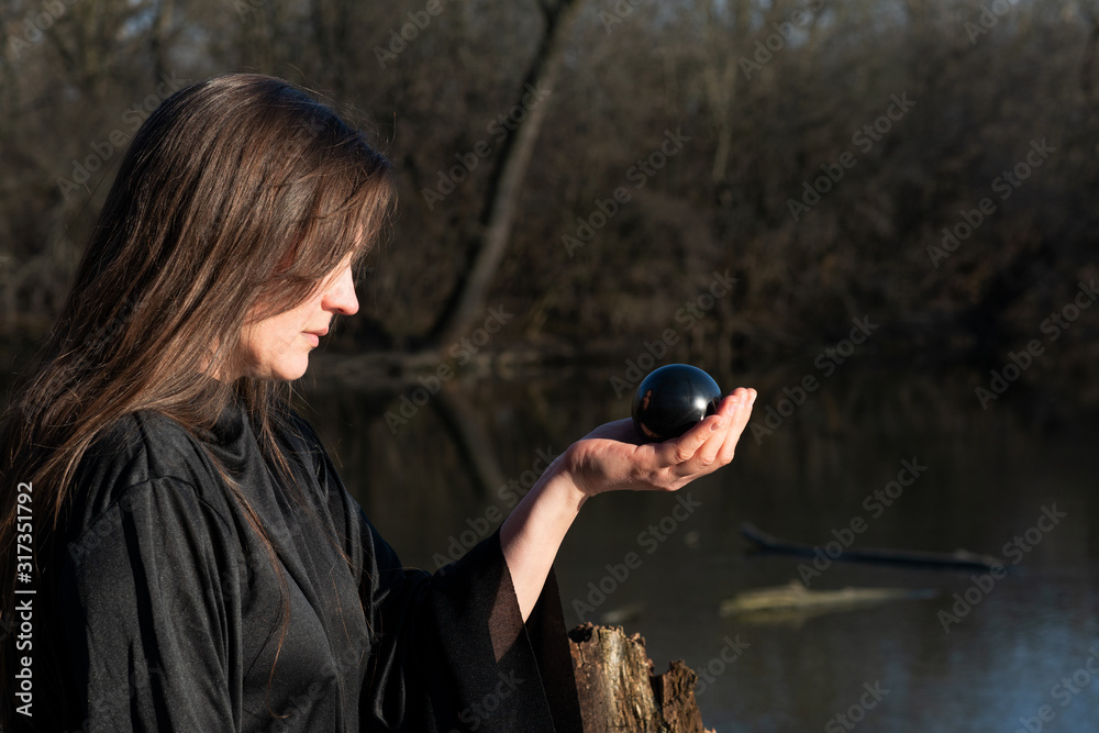 Φωτογραφία Woman with dark long hair in black robes is holding polished Shungite Sphere in her hands in front of the lake
