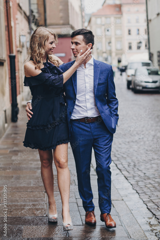 Stylish couple walking together in rainy european city street on background of old architecture. Fashionable man and woman in love enjoying day in city. Traveling together in Europe Stylish couple walking together in rainy european city street on background of old architecture. Fashionable man and woman in love enjoying day in city. Traveling together in Europe