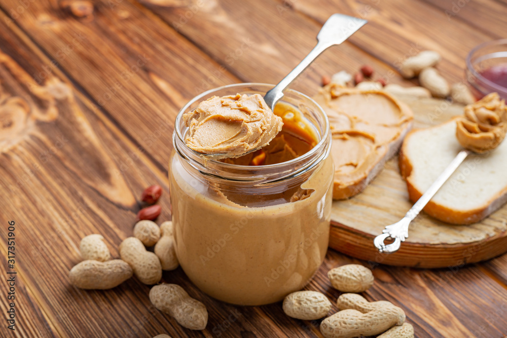 Peanut butter in spoon near creamy peanut paste in open glass jar, slice of peanut butter bread, toast. Peanuts in the peel scattered on the brown wooden table with copy space for cooking breakfast Peanut butter in spoon near creamy peanut paste in open glass jar, slice of peanut butter bread, toast. Peanuts in the peel scattered on the brown wooden table with copy space for cooking breakfast