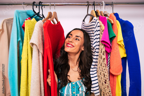 A happy face of shop lady. Close-up photo of a happy girl, who is smiling while sitting inside a wardrobe among multicolored pieces of clothing, touching them with her hands.
