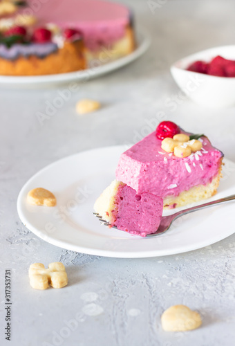 Piece of raspberry cream mousse cake (no baked cheesecake) decorated with fresh raspberries, mini cookies and coconut flakes on light grey background.