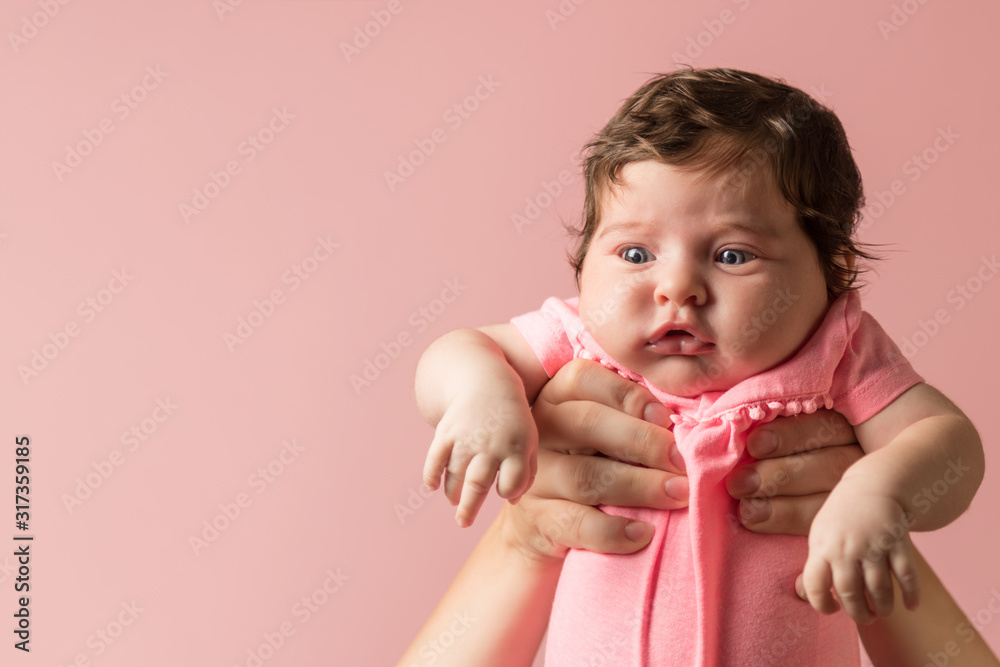 Cute infant in pink jumpsuit in mother hands on light pink background. Maternity concept. Tearfully baby. Copy space - obrazy, fototapety, plakaty 
