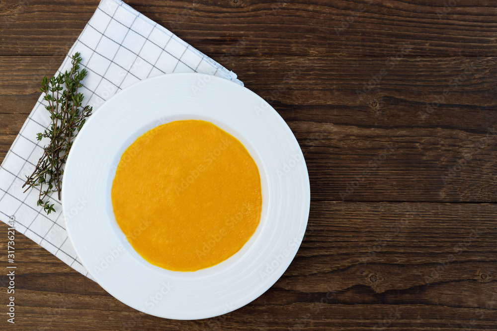 Pumpkin puree soup with a white ceramic plate on a wooden table. Next to a checkered napkin and a bunch of thyme. Copy space Pumpkin puree soup with a white ceramic plate on a wooden table. Next to a checkered napkin and a bunch of thyme. Copy space