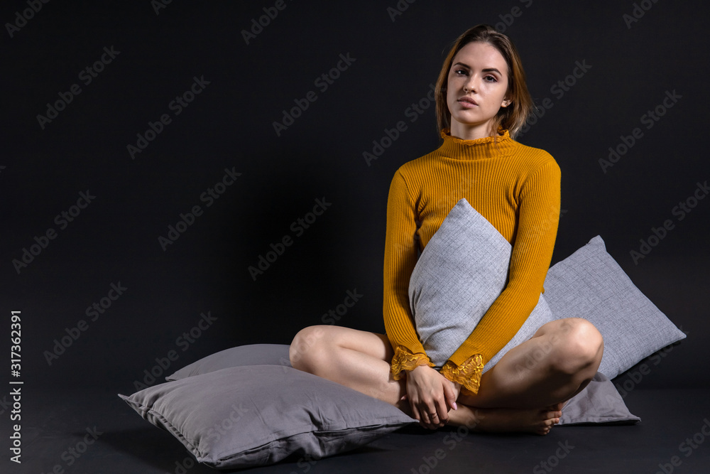 young beautiful girl sitting on the floor among pillows bending legs against a dark background young beautiful girl sitting on the floor among pillows bending legs against a dark background