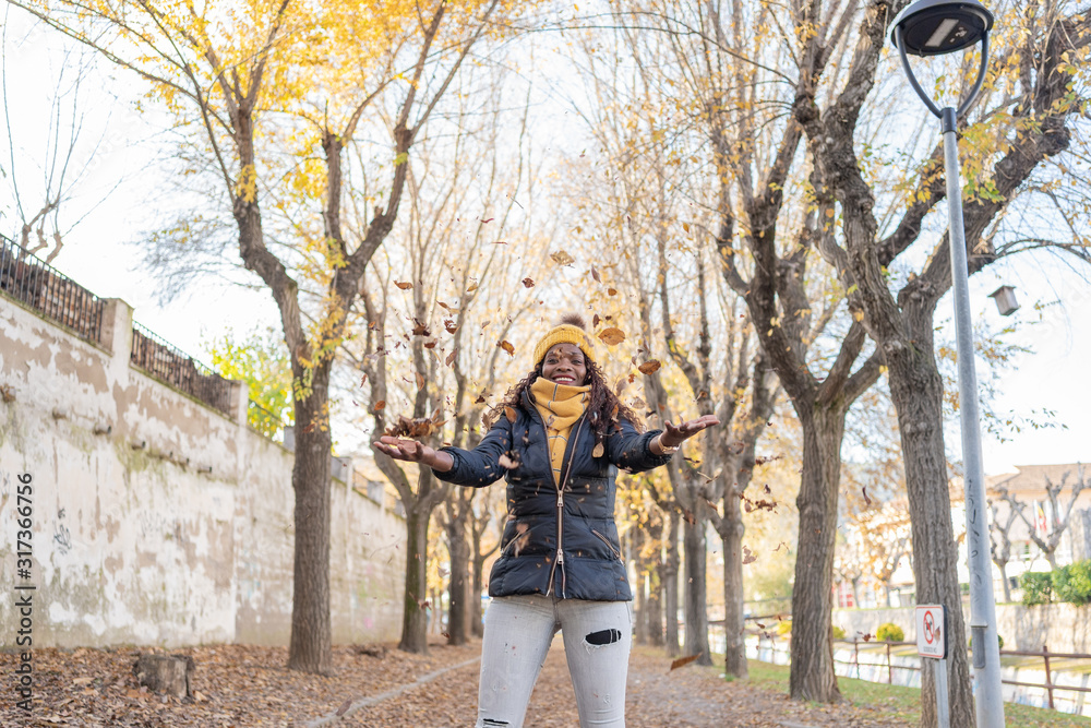 Stylish enthusiastic African American woman in warm jacket merrily throwing dry autumn leaves into air in park