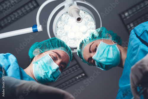 From below female surgeons in medical uniform using professional tools while standing under bright light in operating theater photo