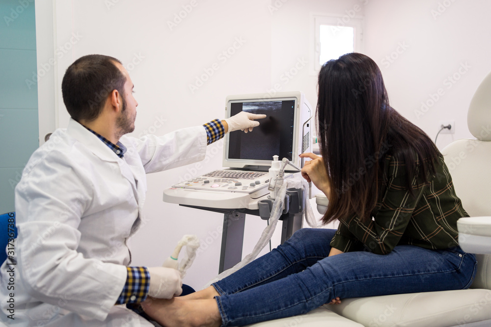 Side view of qualified male podiatry doctor using ultrasound diagnostic equipment and pointing at monitor while examining and consulting  female patient in clinic